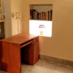 A small room features a wooden desk under a lit shelf holding books and a lamp. To the left, a TV displays a person speaking, with an air conditioner above. To the right, a decorative Buddha artwork is partially visible.